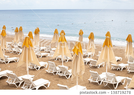Beach scene with empty closed umbrellas and sun loungers on on sea shore Beach scene with empty closed umbrellas and sun loungers on on sea shore 138719278