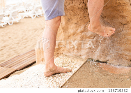 Man washing his feet with shower to clean them from the sand standing on the beach on the seashore. Man washing his feet with shower to clean them from the sand standing on the beach on the seashore. 138719282