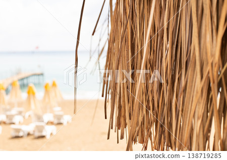 Thatched roof with tropical beach with sand and deck chairs on the sea shore in background. Thatched roof with tropical beach with sand and deck chairs on the sea shore in background. 138719285