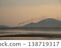 Serene landscape at dawn with factory chimney emitting smoke against backdrop of hills and calm body of water 138719317