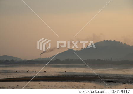 Serene landscape at dawn with factory chimney emitting smoke against backdrop of hills and calm body of water Serene landscape at dawn with factory chimney emitting smoke against backdrop of hills and calm body of water 138719317