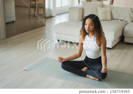 Yoga mindfulness meditation. Young healthy African girl practicing yoga at home. Woman sitting in lotus pose on yoga mat meditating relaxing indoor. Girl doing breathing practice. Yoga at home 138719365