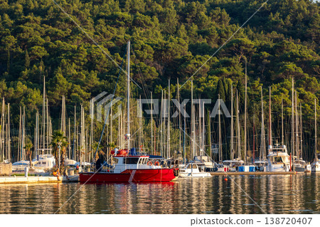 Red fireboat moored in Croatian marina surrounded by sailboat masts, calm water reflections, pine forest hillside. Adriatic harbor scene with nautical atmosphere, summer travel destination europe 138720407
