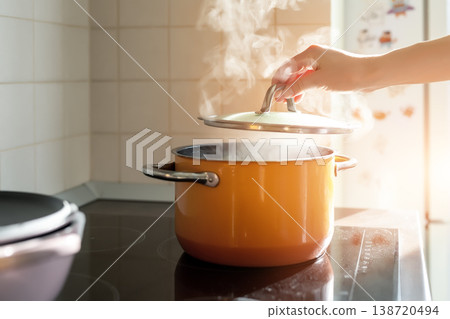 Female hand open lid of enamel steel cooking pan on electric hob with boiling water or soup and scenic vapor steam backlit by warm sunlight at kitchen. Kitchenware utensil and tool at home background Female hand open lid of enamel steel cooking pan on electric hob with boiling water or soup and scenic vapor steam backlit by warm sunlight at kitchen. Kitchenware utensil and tool at home background 138720494