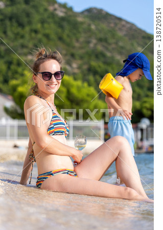 Smiling young adult woman relax bikini sunny beach hold glass wine child play water inflatable arm. Summer vacation lifestyle family carefree leisure mediterranean coast green mountains background Smiling young adult woman relax bikini sunny beach hold glass wine child play water inflatable arm. Summer vacation lifestyle family carefree leisure mediterranean coast green mountains background 138720514
