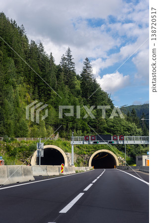 Alpine highway entry twin tunnel portals dense forested hillside transport gantry signals speed restrictions. Scenic European mountain landscape asphalt roadway leading illuminated tunnel passage Alpine highway entry twin tunnel portals dense forested hillside transport gantry signals speed restrictions. Scenic European mountain landscape asphalt roadway leading illuminated tunnel passage 138720517
