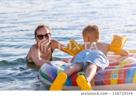 Mother and young boy sit inflatable raft calm sea water marina warm sunset light. Summer vacation moment shows family bonding, happiness, parenting care, childhood joy, and relaxed travel lifestyle Mother and young boy sit inflatable raft calm sea water marina warm sunset light. Summer vacation moment shows family bonding, happiness, parenting care, childhood joy, and relaxed travel lifestyle 138720544