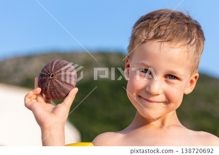Boy holding sea urchin shell found beach sunny summer vacation near Mediterranean coast. Curious child exploring marine nature while enjoying seaside travel, discovery carefree childhood adventure 138720692