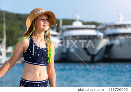 Girl wearing straw hat swimsuit stand sea luxury yachts marina during sunny summer vacation. Travel lifestyle, childhood freedom and seaside tourism atmosphere with blue water and resort background Girl wearing straw hat swimsuit stand sea luxury yachts marina during sunny summer vacation. Travel lifestyle, childhood freedom and seaside tourism atmosphere with blue water and resort background 138720693