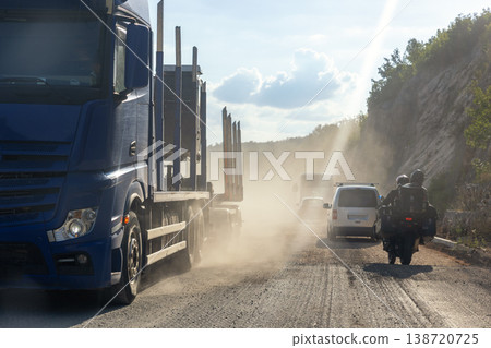 Road construction traffic moving dusty unpaved gravel highway truck, motorcycle vehicles sunlight rocky hillside. Transport scene heavy traffic dirt surface haze infrastructure repair rural landscape 138720725