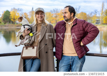 Happy Adult Couple With Small Dog Standing Near Autumn Lake Outdoor Portrait Happy Adult Couple With Small Dog Standing Near Autumn Lake Outdoor Portrait 138721015