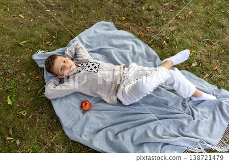 Young boy relaxing on a soft blanket in the park with a fresh red apple beside him. Young boy relaxing on a soft blanket in the park with a fresh red apple beside him. 138721479