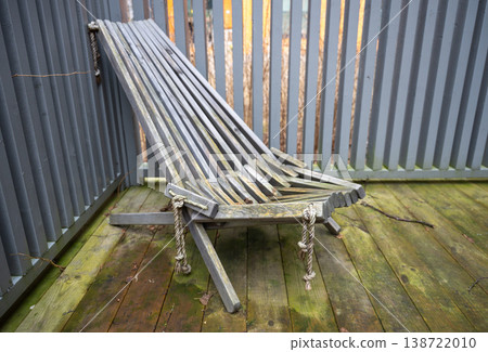 Folded wooden deck chair on weathered terrace with gray fence and rustic outdoor texture 138722010