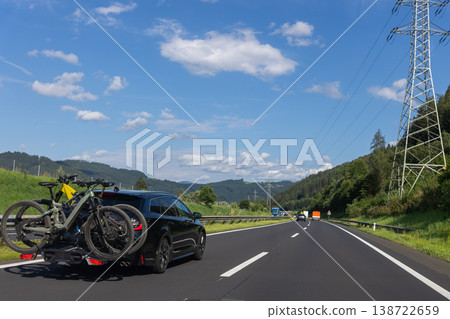 Car bicycles on rear carrier driving along mountain highway forest valley blue sky clouds sunny day. Summer road trip scene travel cycling adventure transport mobility countryside tourism 138722659