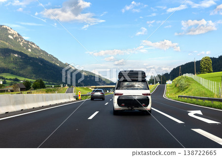 Family van with roofbox driving along alpine highway between green mountains blue sky. Road trip travel scene showing summer mobility tourism freedom adventure scenic countryside transport route Family van with roofbox driving along alpine highway between green mountains blue sky. Road trip travel scene showing summer mobility tourism freedom adventure scenic countryside transport route 138722665