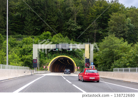 Cars driving tunnel entrance highway surrounded green forest hills daylight conditions. Road infrastructure signals speed limit signs barriers guides traffic flow into illuminated tunnel passage Cars driving tunnel entrance highway surrounded green forest hills daylight conditions. Road infrastructure signals speed limit signs barriers guides traffic flow into illuminated tunnel passage 138722680