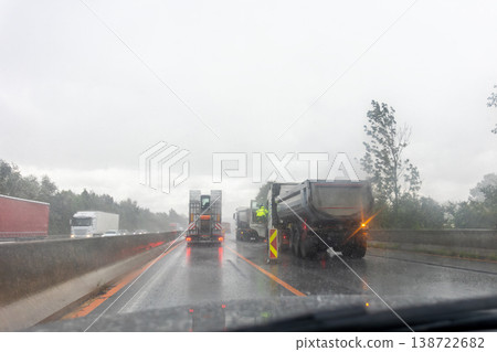 Driver POV rain highway roadworks trucks workers wear safety gear traffic poor visibility wet asphalt conditions. Vehicles move slowly construction site zone warning signs barriers reflective lights 138722682