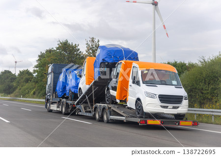 Truck carries multiple double cabin vans covered colorful tarps highway under cloudy sky wind turbines background. Commercial transport scene logistics delivery service renewable energy landscape Truck carries multiple double cabin vans covered colorful tarps highway under cloudy sky wind turbines background. Commercial transport scene logistics delivery service renewable energy landscape 138722695