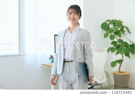 A young Japanese female businessperson in a light gray suit stands in an office, holding files and smiling. A young Japanese female businessperson in a light gray suit stands in an office, holding files and smiling. 138723485