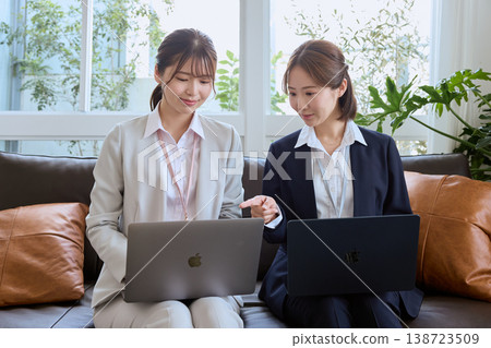 兩位年輕的職業婦女正在辦公室的沙發上，一邊看著筆記型電腦一邊開會討論工作。 138723509