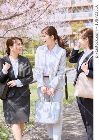 Three female new employees, dressed in suits and smiling, walk under a row of cherry trees—an image of a spring induction ceremony. Three female new employees, dressed in suits and smiling, walk under a row of cherry trees—an image of a spring induction ceremony. 138723516