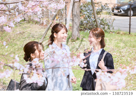 Three new female employees smiling and chatting under cherry blossoms – a fresh image of a springtime office district. 138723519