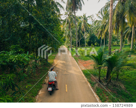 Traveler riding a scooter on a quiet road through a tropical palm forest. Traveler riding a scooter on a quiet road through a tropical palm forest. 138724989