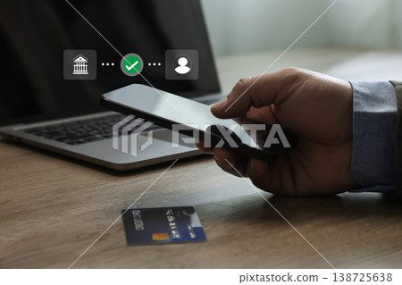 Hand holding a smartphone near a laptop with a credit card on the table, showing digital payment authentication and financial transaction process in a modern indoor environment. 138725638