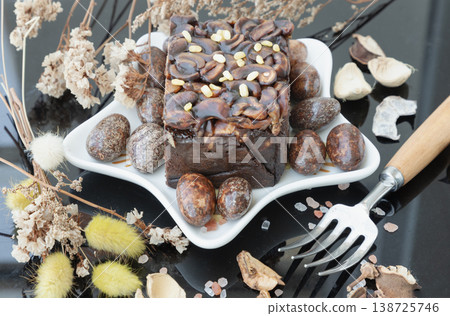 Dark toffee cheesecake topped with cashews and toasted mung beans, served on a star-shaped plate with chocolate-coated almonds and a side fork. The aesthetic is completed by a rustic backdrop of dried 138725746