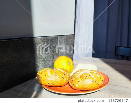 Two seeded bread rolls are placed on an orange plate on a wooden table. A ripe apple rests nearby. Breakfast in cafe 138726347