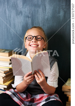 Girl reading book, student and chalkboard for education, language learning and knowledge in classroom. Smart kid or child with glasses and happy for school library, fiction and literature in portrait Girl reading book, student and chalkboard for education, language learning and knowledge in classroom. Smart kid or child with glasses and happy for school library, fiction and literature in portrait 138727587