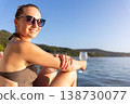 Smiling woman in sunglasses relaxes near calm water holding glass during warm sunset. Natural beauty, summer vacation mood, peaceful landscape freedom leisure serene travel atmosphere by quiet shore 138730077
