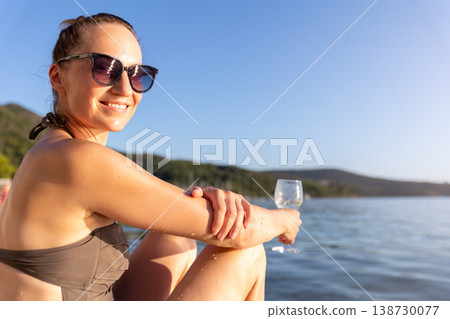 Smiling woman in sunglasses relaxes near calm water holding glass during warm sunset. Natural beauty, summer vacation mood, peaceful landscape freedom leisure serene travel atmosphere by quiet shore 138730077