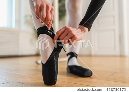 Ballerina puts on pointe shoes on leg in dance class. Classical ballet dancer woman in dance studio. Ballerina putting on tying classical pointe shoes for dance training. Ballet school concept 138730173