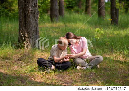 mother and son are sitting side by side in a forest clearing and looking at something in their palms 138730547
