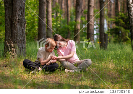 mother and son are sitting side by side in a forest clearing and looking at something in their palms mother and son are sitting side by side in a forest clearing and looking at something in their palms 138730548