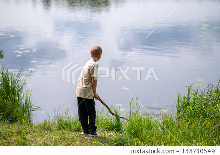 Boy standing on lakeside with a long stick, playing near the water in nature on a summer day Boy standing on lakeside with a long stick, playing near the water in nature on a summer day 138730549