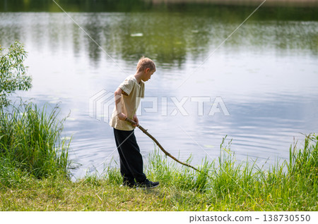 Boy standing on lakeside with a long stick, playing near the water in nature on a summer day Boy standing on lakeside with a long stick, playing near the water in nature on a summer day 138730550