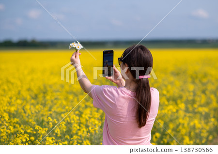 Woman taking photo of daisies on smartphone in blooming yellow rapeseed field, summer flower photography in nature Woman taking photo of daisies on smartphone in blooming yellow rapeseed field, summer flower photography in nature 138730566