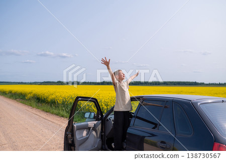 Happy boy standing with raised arms on country road in rapeseed field near car on sunny summer day, joyful outdoor adventure Happy boy standing with raised arms on country road in rapeseed field near car on sunny summer day, joyful outdoor adventure 138730567