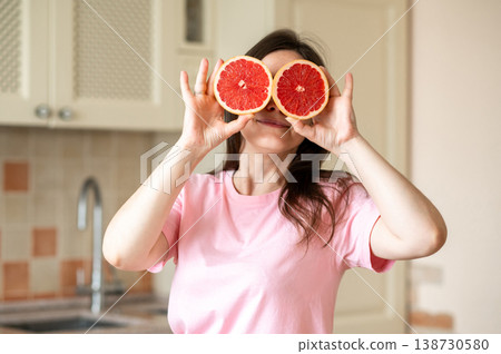 Happy woman holding grapefruit halves over her eyes while standing in kitchen with fresh fruits Happy woman holding grapefruit halves over her eyes while standing in kitchen with fresh fruits 138730580
