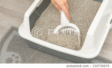 Hand using a scoop to clean cat litter in a gray litter box placed on a textured mat with a wooden floor background 138730870