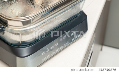 Glass container with lid on induction cooktop showing steam condensation and food inside during cooking. Home kitchen steaming process, covered dish heating, appliance usage, food preparation. 138730876