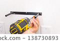 Mans hand holding yellow cordless drill fastening black metal bracket to white wall with screw. Home improvement, securing shelf support, DIY mounting, tool in use. 138730893