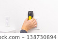Mans hand pressing yellow stud finder on white wall beside power outlet. Home improvement, final wall scan, DIY confirmation, tool application. 138730894
