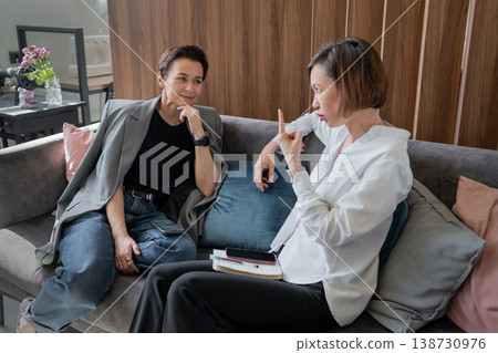 Businesswomen conversing with hand gestures while seated on sofa in stylish office interior. Conversation, gestures, office, teamwork, business, colleagues. 138730976