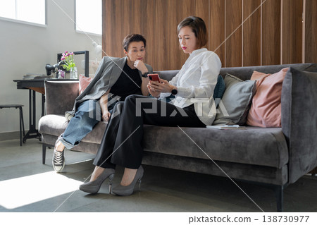 Two coworkers communicating and taking notes with pen on couch in bright workplace setting. Communication, notes, coworkers, teamwork, office. Two coworkers communicating and taking notes with pen on couch in bright workplace setting. Communication, notes, coworkers, teamwork, office. 138730977