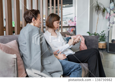 Two professional women talking and reviewing notes together on sofa in creative office. Discussion, notes, business, colleagues, businesswomen, teamwork. 138730982