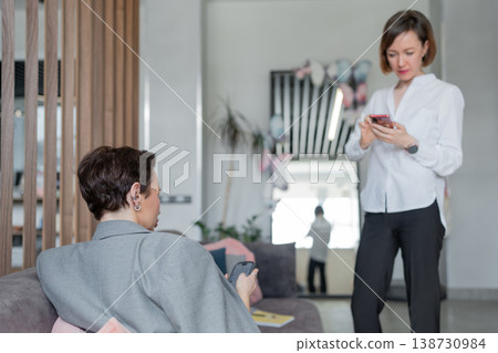 Businesswomen, one standing and one sitting, discussing notes together in stylish modern workspace. Discussion, notes, businesswomen, teamwork, office. 138730984