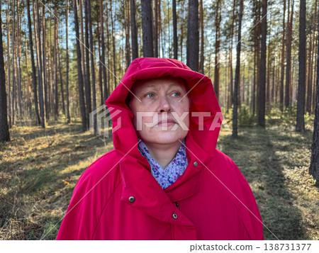 A woman stands in the forest in a red jacket and hood, thoughtfully looking at the surroundings, walking in the fresh air 138731377